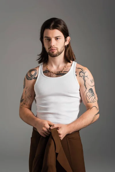 Tattooed long haired man holding jacket standing isolated on grey