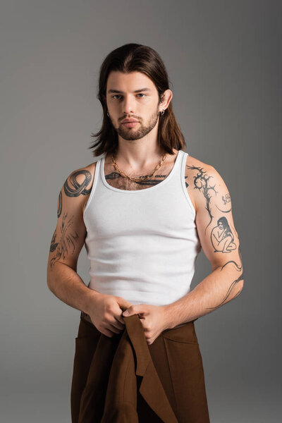 Tattooed long haired man holding jacket standing isolated on grey 