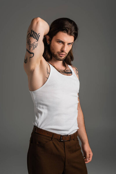 Muscular and tattooed model in white tank top looking at camera isolated on grey 