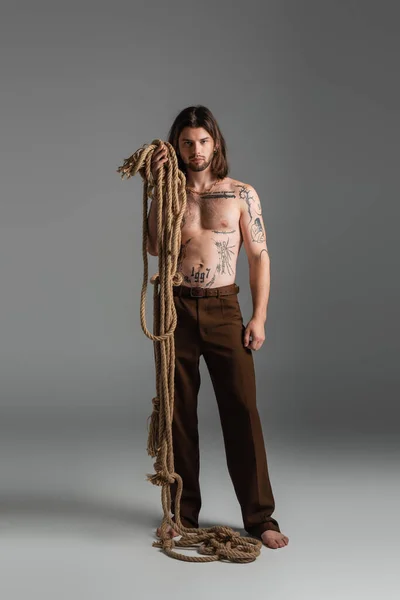 Barefoot and tattooed model holding rope on grey background