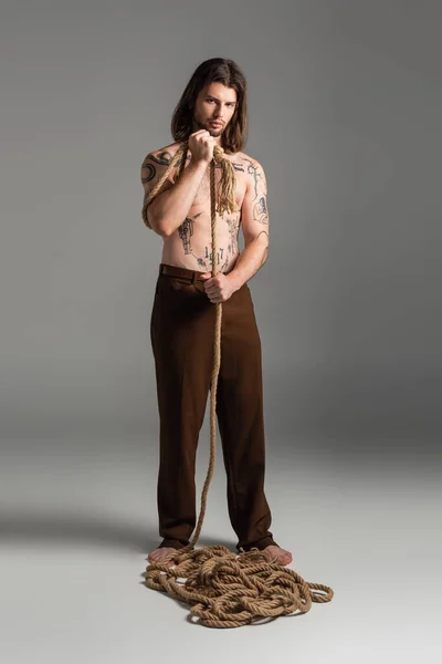 Full length of tattooed man in pants holding rope on grey background