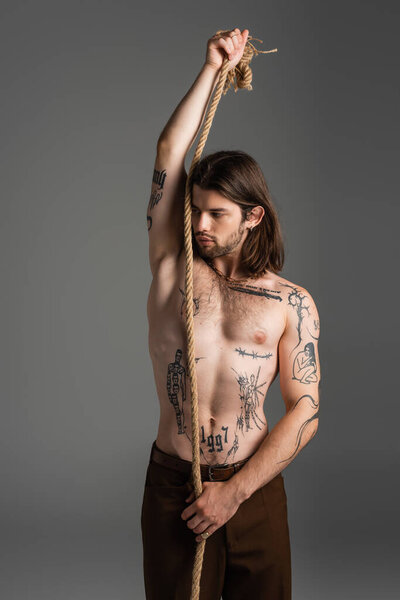 Shirtless tattooed man holding rope and looking away isolated on grey 