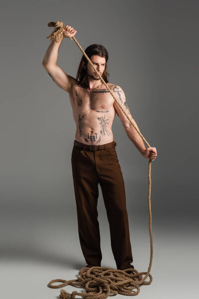 Full length of tattooed model in pants holding rope on grey background 