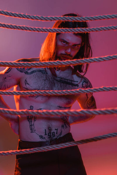 Young tattooed man standing near ropes on purple background with red light 