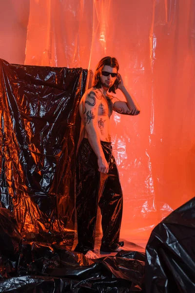 Long haired and tattooed man in latex pants standing on cellophane