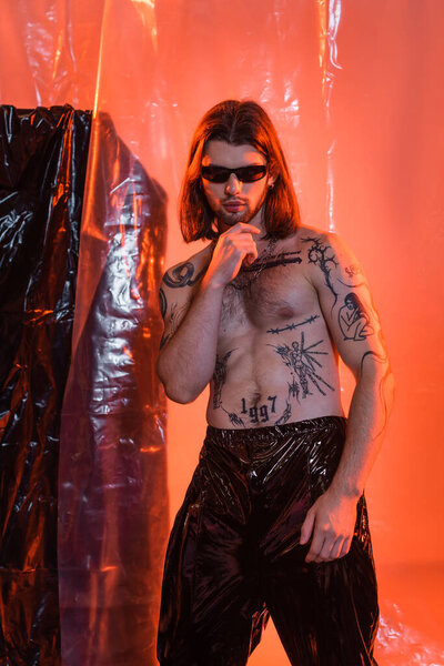 Tattooed man in sunglasses and latex pants posing near polyethylene