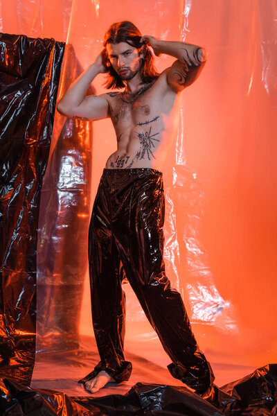 Full length of tattooed man in latex pants touching hair near cellophane 