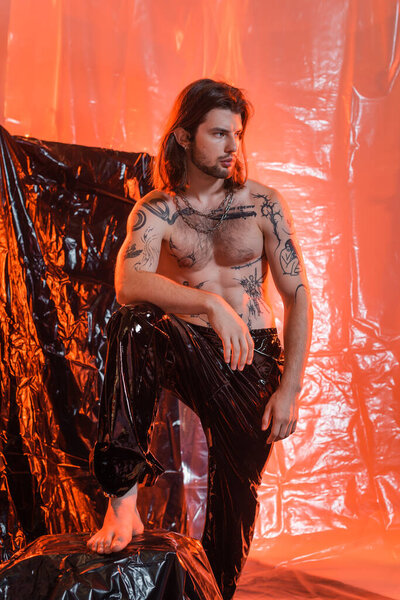Sexy tattooed man in latex pants standing near cellophane 