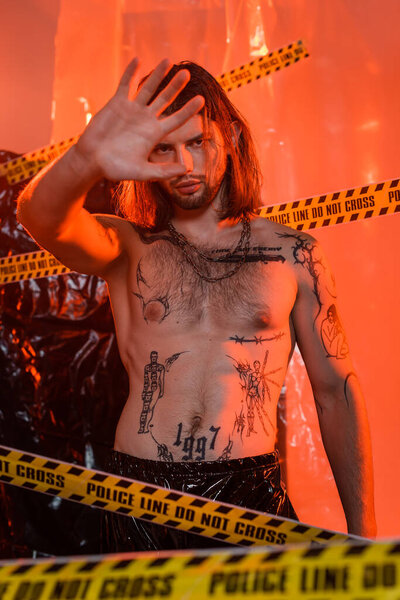 Shirtless tattooed man showing stop gesture near police line and polyethylene at background 