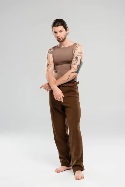 Full length of young barefoot and tattooed model looking at camera on grey background