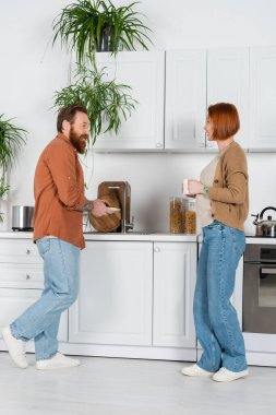 Side view of man washing plate near redhead wife with cup at home  clipart