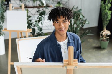 Gülümseyen Afrikalı Amerikalı sanatçı boya fırçası tutuyor ve stüdyoda tuvalin yanındaki kameraya bakıyor. 