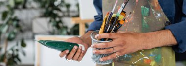 Önlüklü Afro-Amerikan ressamın elinde fırça, boya ve afiş tutarken çekilmiş görüntüsü. 