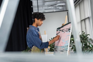 Side view of young african american artist in apron painting on canvas  clipart