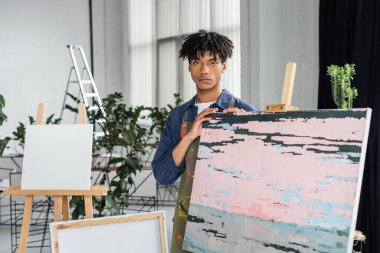 Önlüklü genç Afrikalı Amerikalı sanatçı atölyenin yakınındaki resim makinesine bakıyor. 