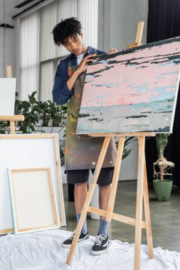 Önlüklü Afrikalı Amerikalı ressam tuvaldeki soyut resme bakıyor. 