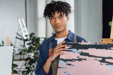 Kirli önlüklü Afrikalı Amerikalı ressamın portresi stüdyonun yakınındaki kameraya bakıyor. 