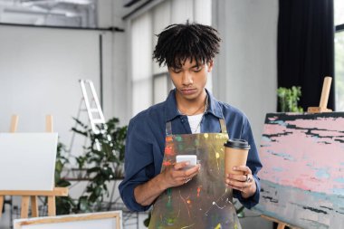 Önlüklü genç Afrikalı Amerikalı sanatçı elinde kağıt bardakla atölyede akıllı telefon kullanıyor. 