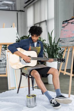 Önlüklü Afrikalı Amerikalı sanatçı stüdyoda resim ve tuvallerin yanında akustik gitar çalıyor. 