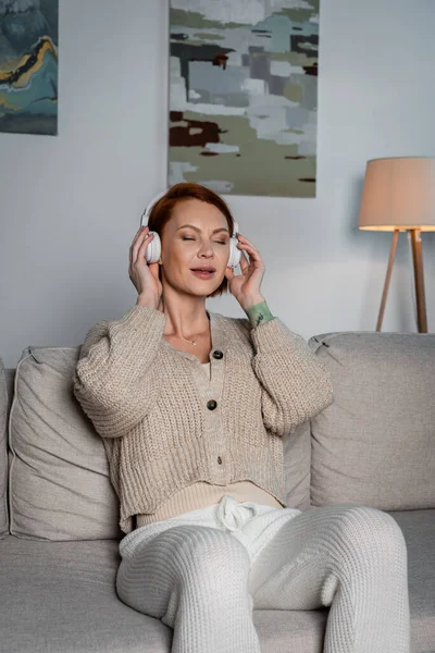 Pleased woman in wireless headphones listening music on couch