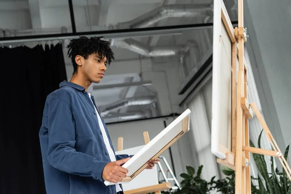 stock image Low angle view of african american artist holding canvas near easel in studio 