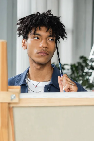 stock image Thoughtful african american artist holding paintbrush near blurred canvas in studio 