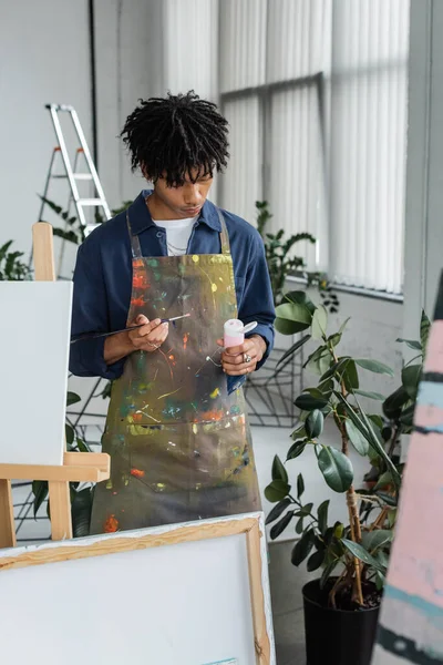 stock image African american artist in apron holding paintbrush and paint near canvases in workshop 