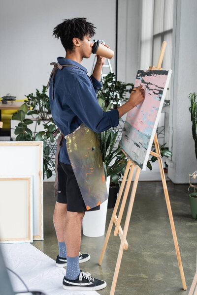 Side view of young african american artist drinking coffee and painting on canvas in studio 