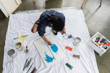 Overhead view of african american artist painting on canvas near paints on floor in studio  clipart