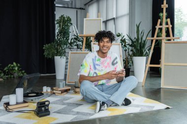 Smiling african american artist using smartphone near vintage camera and record player in workshop  clipart