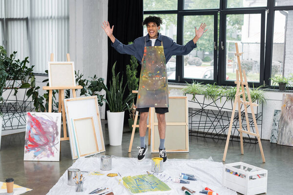 Cheerful african american artist in apron jumping near drawings in studio 