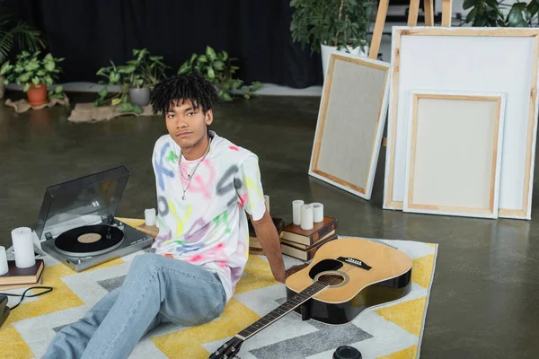 Stock image African american artist looking at camera near acoustic guitar and canvases in workshop 