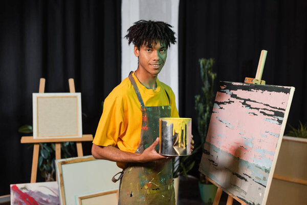 Smiling african american artist holding jar with paint near drawing in workshop 