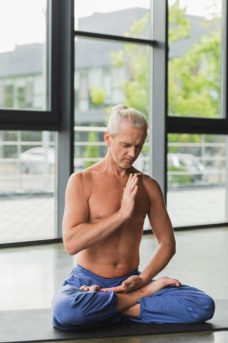 Kapalı gözlü bir adam stüdyoda yoga minderinde poz veriyor. 