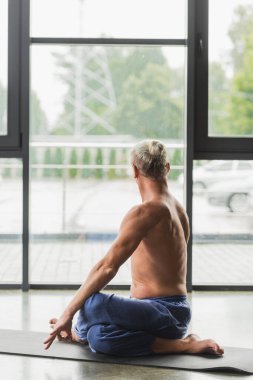 Mavi pantolonlu bir adam yoga pozisyonunda stüdyoda minder üzerinde oturuyor. 