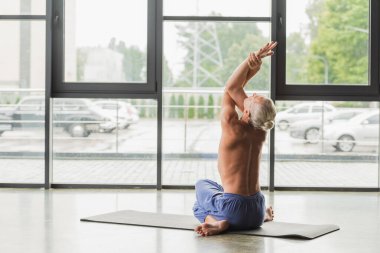 Mavi pantolonlu tişörtsüz adam yoga pozisyonunda oturuyor ve gerinirken kollarını kaldırıyor. 