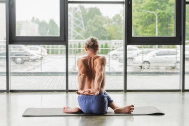 back view of flexible man in blue pants sitting in twisting yoga pose on mat in studio  clipart