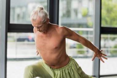 shirtless man in green pants practicing crescent lunge in yoga studio  clipart