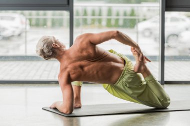 grey haired man doing supine yoga pose while stretching back on yoga mat in studio  clipart