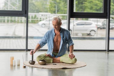 Gri saçlı adam, yoga stüdyosunda Tibet müzik kasesinde poz verirken poz veriyor. 