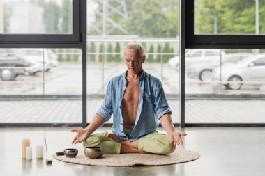 Gri saçlı adam, yoga stüdyosundaki Tibet şarkı kaselerinde meditasyon yaparken nilüferde poz veriyor. 