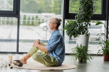Tibet şarkı kaseleri ve tütsü çubukları yanında meditasyon yapan gri saçlı adamın yoga stüdyosunda yan görünüşü. 