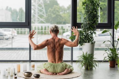 Gömleksiz bir adamın gök gürültüsü yoga pozisyonunda Tibet şarkı kaselerinin yanında otururken el kol hareketi yapması. 