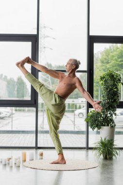 Yeşil pantolonlu üstsüz adam yoga stüdyosunda mumların yanında el pençe divan duruyor. 