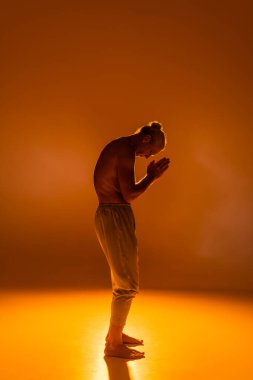 full length of shirtless man in pants practicing yoga with praying hands on orange background  clipart