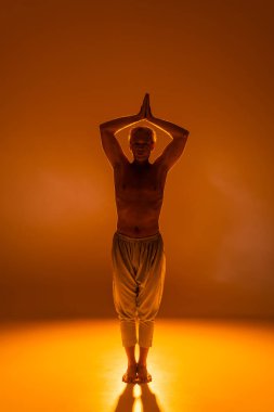 full length of shirtless man in pants standing in warrior pose on orange background  clipart