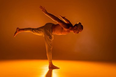 Pantolonlu üstsüz bir adam turuncu üzerinde savaşçı yoga pozu veriyor. 