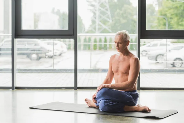 barefoot man in blue pants sitting in twisting yoga pose on mat