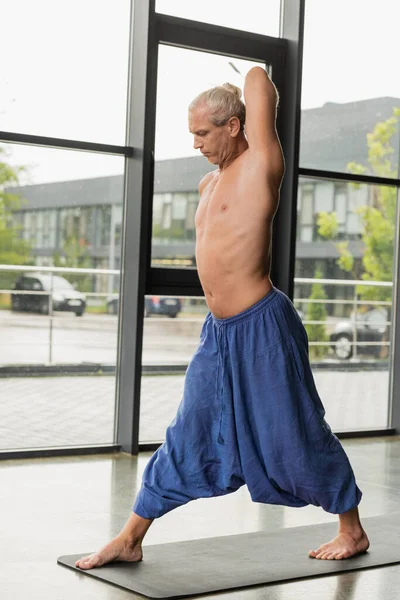 grey haired man in blue pants practicing yoga on mat in studio