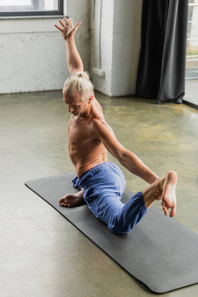 grey haired man doing half pigeon pose with raised hand on yoga mat in studio 